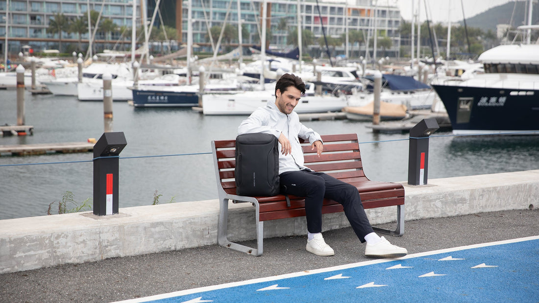 Man with Mark Ryden anti-theft backpack sits on marina bench near boats