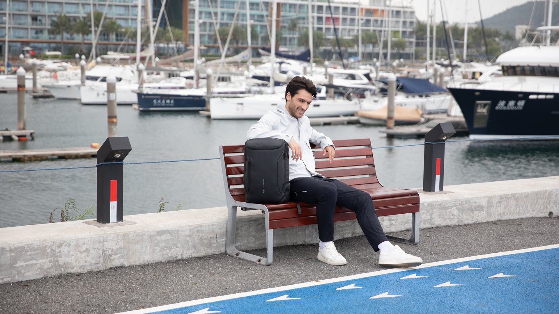 Man with Mark Ryden anti-theft backpack sits on marina bench near boats