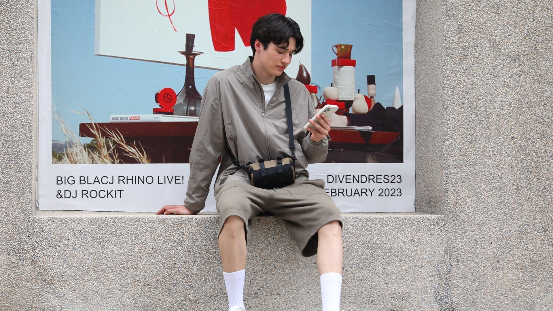 Traveler in gray outfit with Mark Ryden crossbody bag using phone near Lyon travel poster