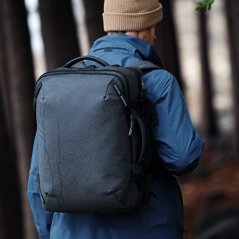 Dark gray Mark Ryden Infinitely multi-functional large capacity backpack with water-resistant material, worn by a person in a blue jacket and beige beanie outdoors among trees.