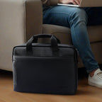 Black MarkRyden Tech Commuter Computer Briefcase 14/15.6 inch with dual padded handles, textured exterior, placed on wooden floor with laptop user in background.