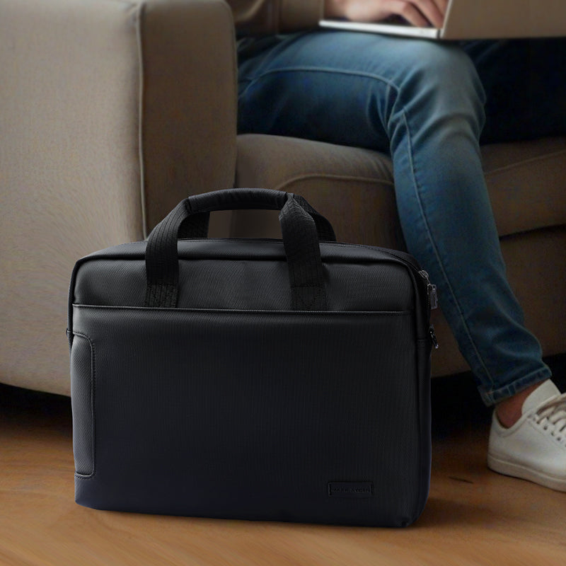 Black MarkRyden Tech Commuter Computer Briefcase 14/15.6 inch with dual padded handles, textured exterior, placed on wooden floor with laptop user in background.