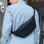Black sleek crossbody bag with zippered front pocket, worn by a person in a light blue coat, perfect for daily fashion use.