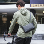 Black lightweight waterproof crossbody bag worn by a man in a green jacket while riding a bike near a café.