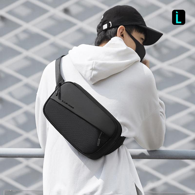Mark Ryden CrossWear black waterproof crossbody bag with multiple compartments and front zip pocket, worn by person in white hoodie and black cap against grid background.
