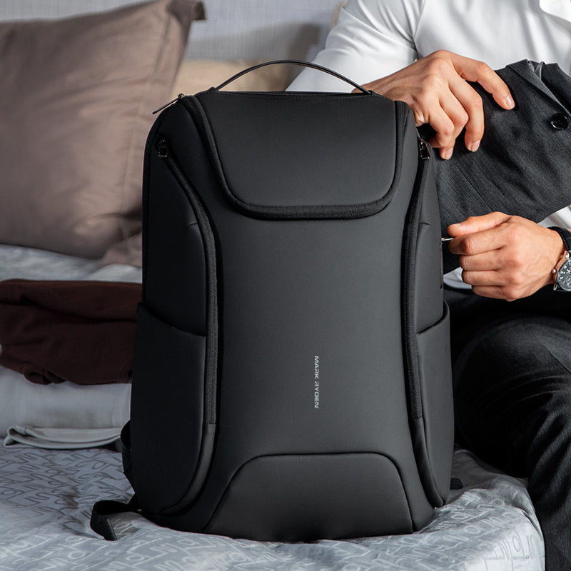 Black Mark Ryden Polycom 2nd waterproof backpack with built-in USB, displayed on a bed with travel clothing nearby.